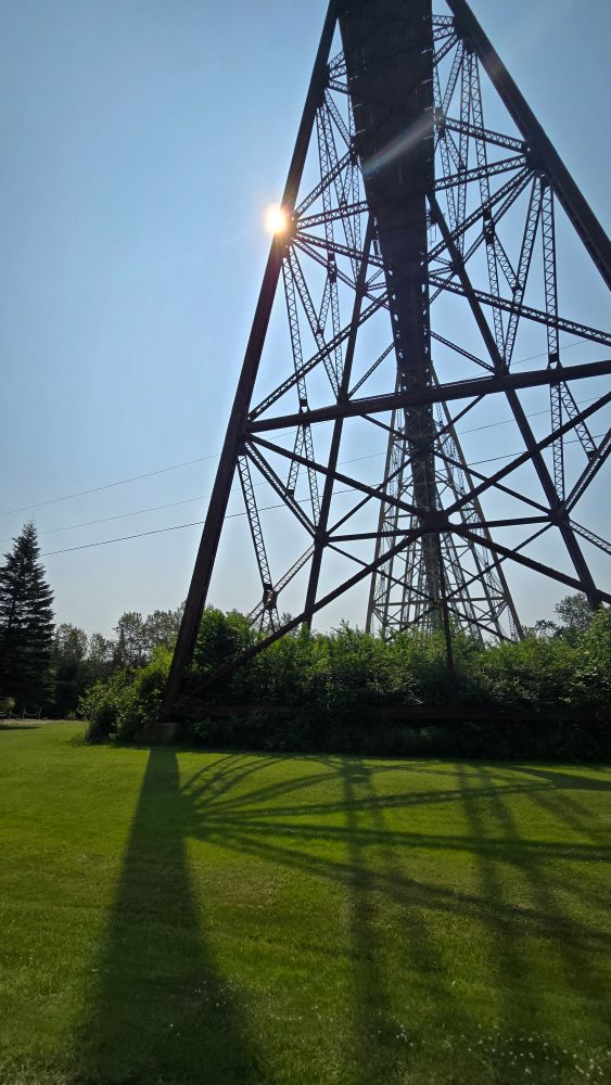A picture of the sun shining on a railway trestle. Taken from below the train bridge, the sun is casting a shadow of the tressle on the grass. 