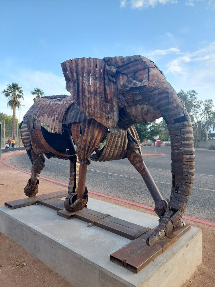 A photo of a large statue of an elephant made from rusty reclaimed pieces of piping and corrugated metal. The pipe legs are disproportionately skinny, giving a skeletal kind of look, and gaps between metal pieces in the body and head give the impression of rotting.