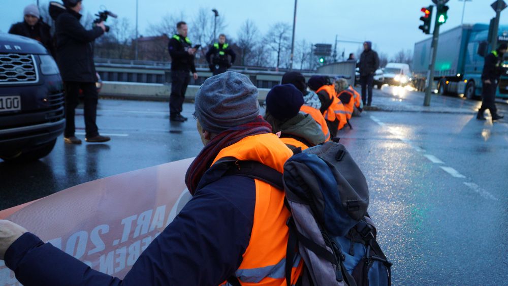 Reihe an Menschen in orangenen Warnwesten sitzt auf nasser Fahrbahn, ein Auto hält davor, Presse filmt, Polizisten warten. Von seitlich hinten fotografiert.