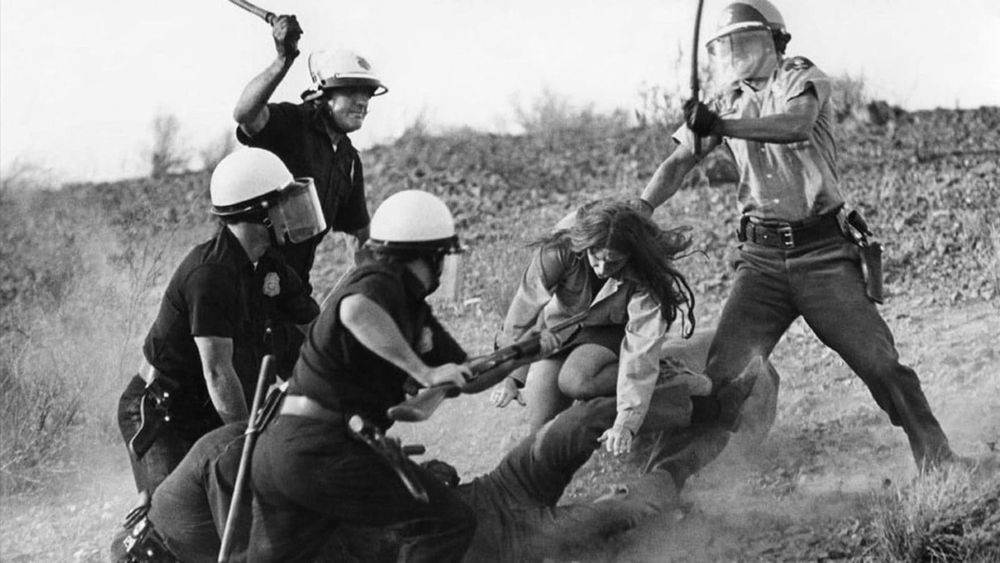 Four cops are beating a person who is on the ground of a desert. There is a woman who is trying to help him.