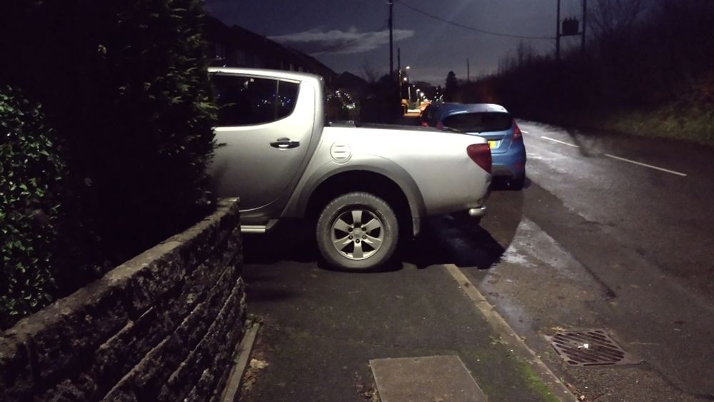 View along a pavement at night, the pavement completely blocked by the rear half of a double cab pickup, half way into a house drive, overhanging into the road.