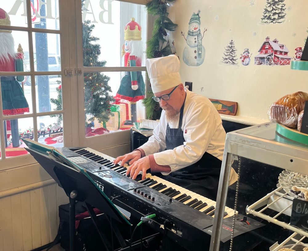 A man in a chef’s hat oak keyboard, surrounded by baked goods and winter holiday decorations. 