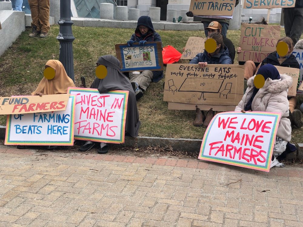 Several women in traditional dress sit holding signs that say "The heart of farming beats here" - "Invest in Maine Farms" - We LOVE Maine Farmers" - "Everyone Eats, empty plate, empty future" - SAVE BEES, NOT Billionaires" & "Cut Flowers, Not Funds"