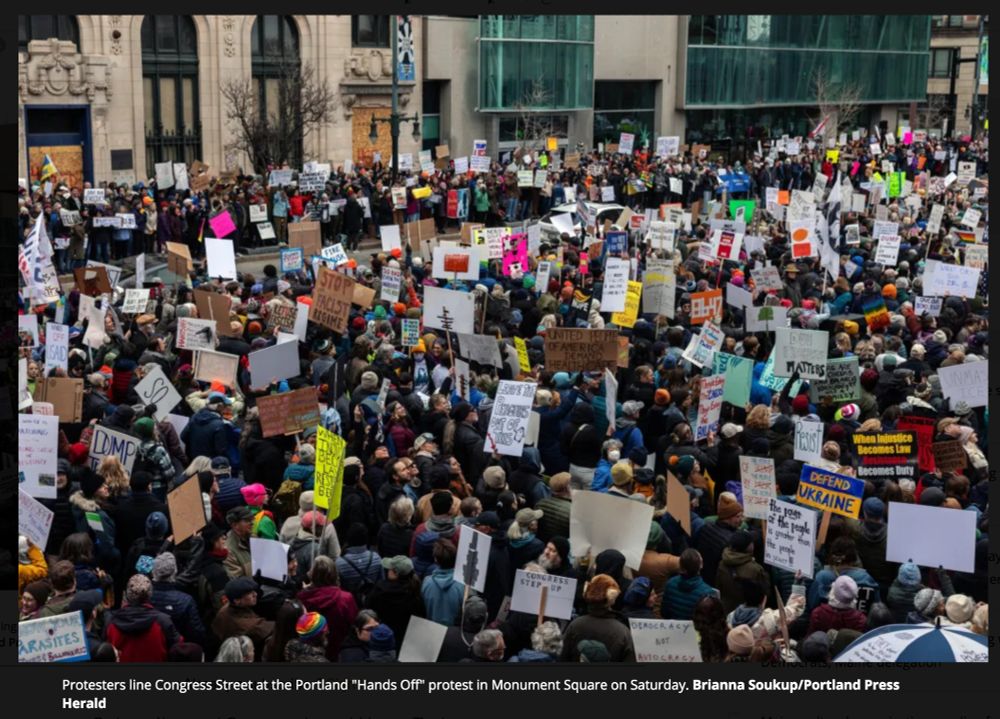 Throngs of protesters holding signs line both sides of a street in a downtown scene.