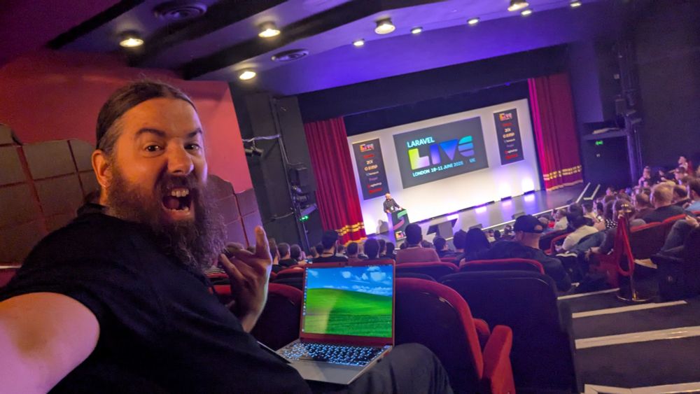 A selfie of me, to the left, with a Framework laptop with the Windows XP "Bliss" background on my lap. Behind and centre frame is the stage at the Shaw Theatre, with the Laravel Live conference branding displayed on a large projection screen, with a list of the conference sponsors either side. There is a lectern on the stage, behind which Jon the conference organiser is stood, about to make the welcome address.