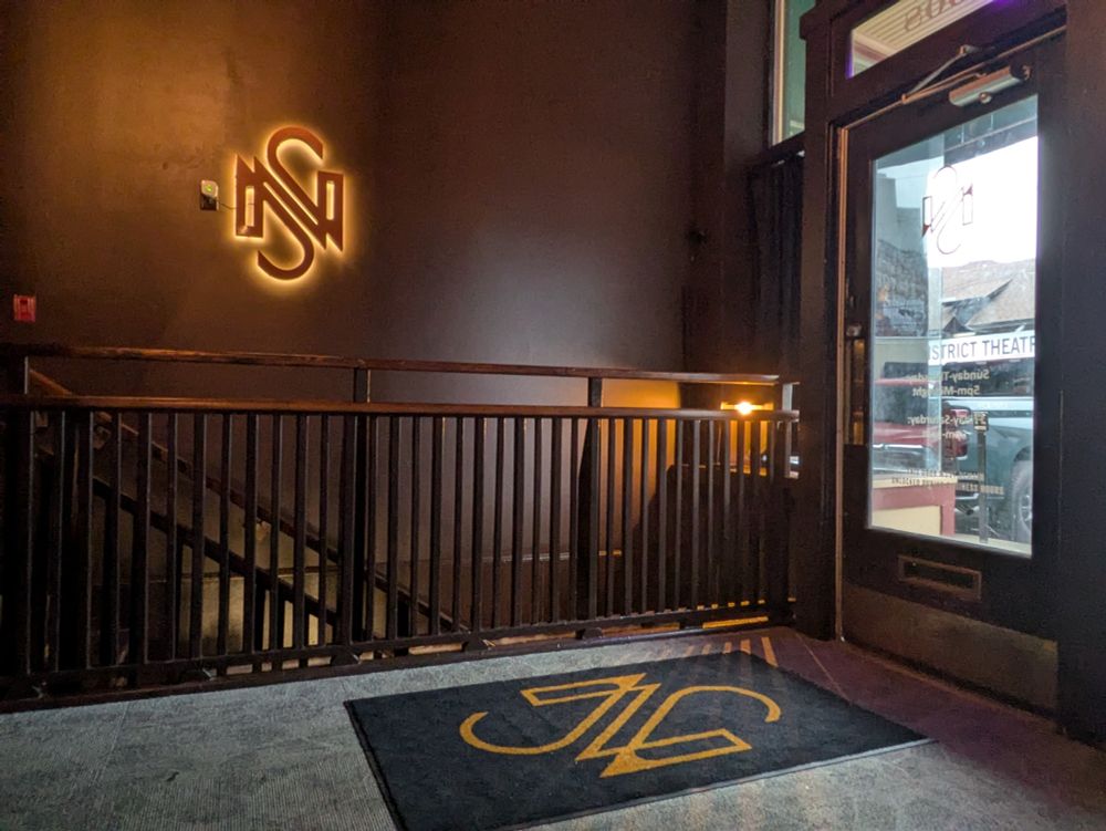 The lobby of Nowhere Special, a basement bar in Indianapolis. It's early evening and the sun coming through the window decorated the N/S logo on the carpet. On the brown wall, a backlit logo glows. 
