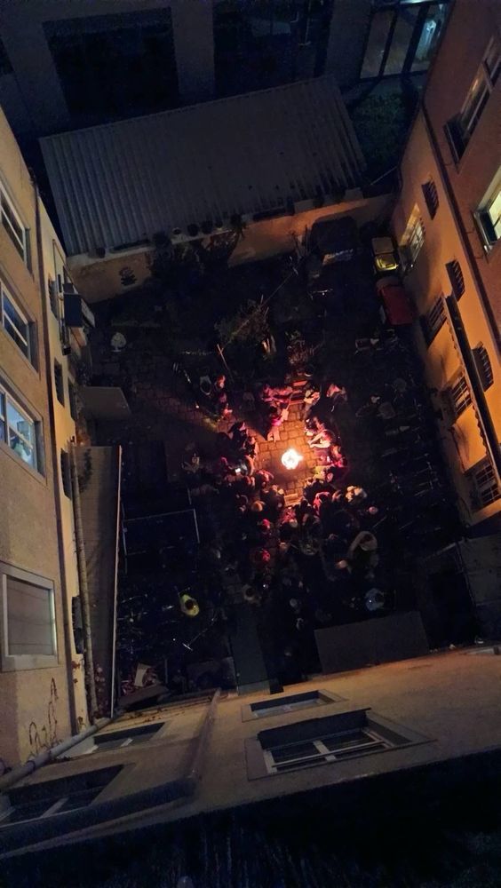 Das Bild zeigt aus der Vogelperspektive den Innenhof des Hausprojekts Willy*Fred. Das Foto wurde in der Nacht aufgenommen und es ist ein Lagerfeuer zu sehen, um das sich viele Menschen tummeln.