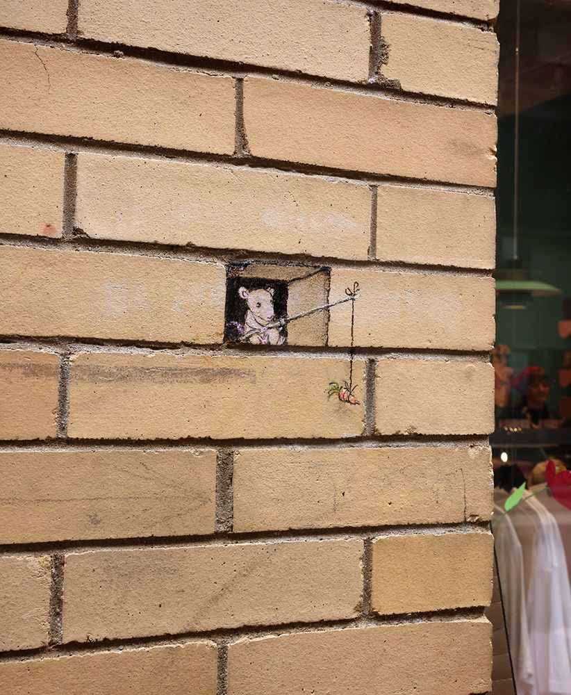 chalk drawing of a mouse sitting in a gap between bricks in a wall, dangling a carrot from a stick