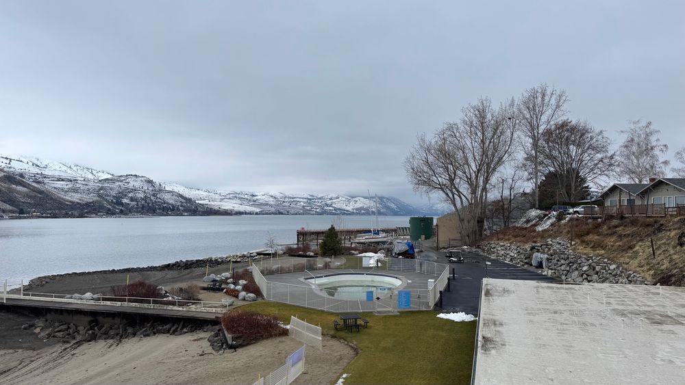 Overlooking a grass area with a hot tub. There is a lake in the background with snowy hills behind that. There are low clouds.