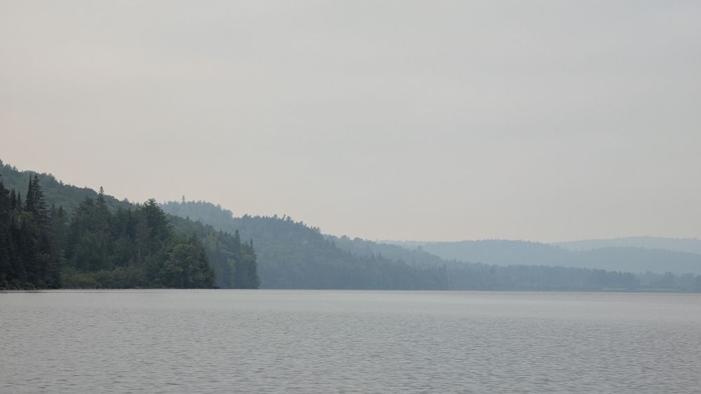 A calm lake, with forested hills in the distance. A haze makes the hills fade to light blue in the distance. The sky is grey.
