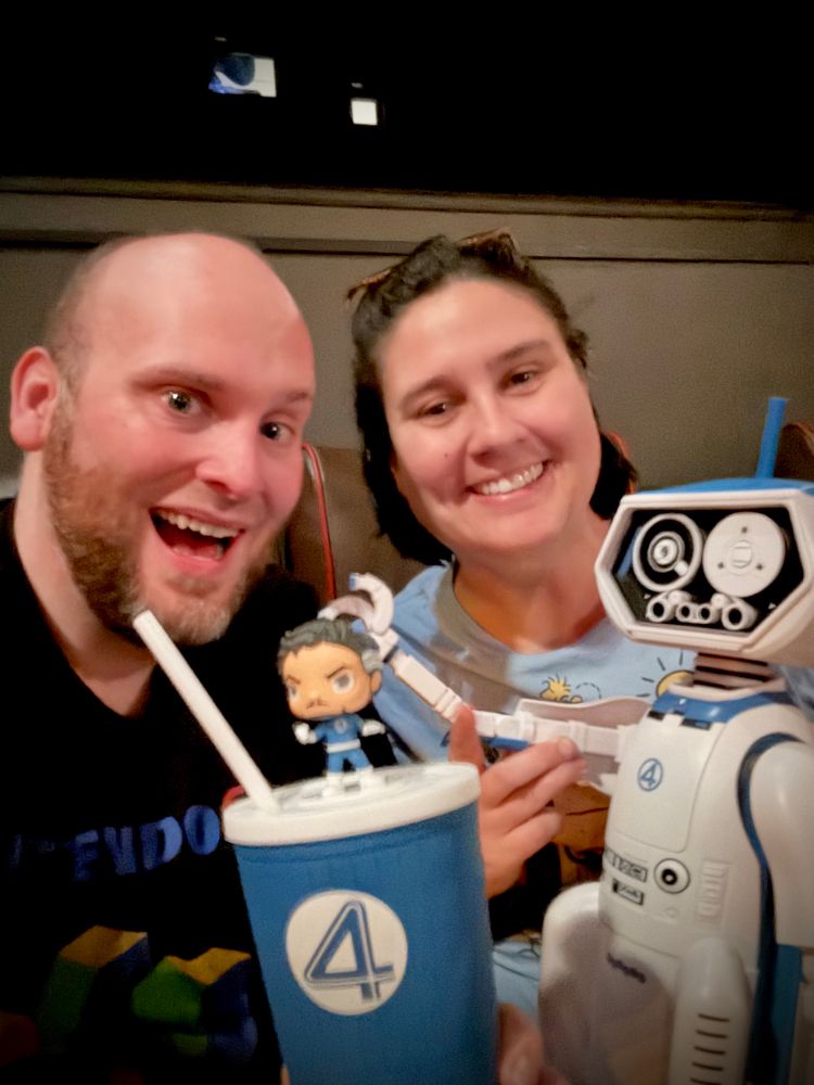 Matt and Maggie are smiling in a movie theater, holding themed merchandise from the new Fantastic Four movie. In front of them is a blue cup with the Fantastic Four “4” logo, topped with a small figure of Mr. Fantastic. Next to them is a white and blue HERBIE robot popcorn bucket, also branded with the “4” logo.