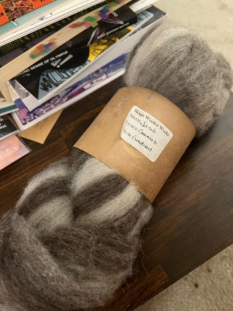 Gray (light and dark together) wool twisted into a skein with a brown paper band around it. The band has a sticker that says “breed: Jacob 
Source: Camano Is. 
Color: gradient”