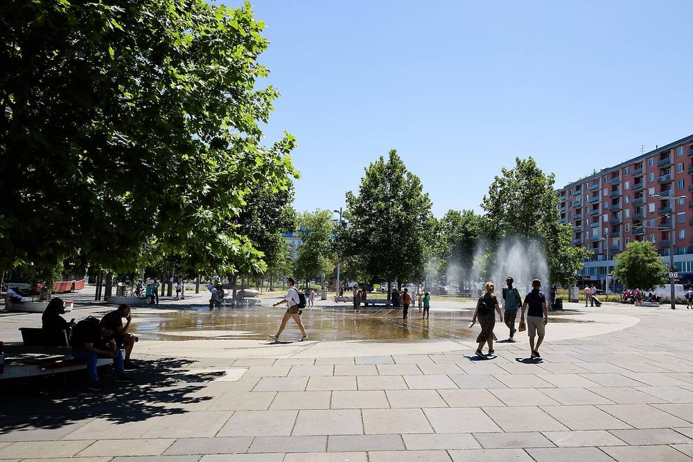 Menschen gehen über den Praterstern, ein Platz mit Betonfliesen, in der Mitte ein Springbrunnen. Am Rand sitzen Bäume im Schatten unter einem Baum. Im Hintergrund noch mehr Bäume