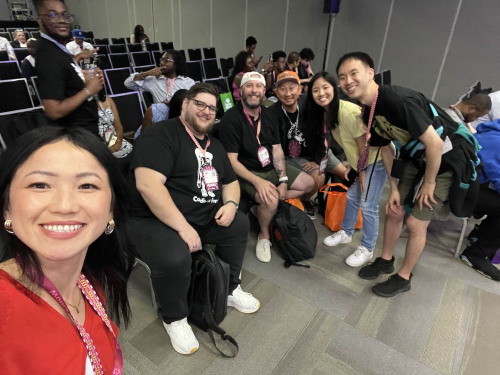 7 women and men sitting on chairs or standing in a room full of chairs
