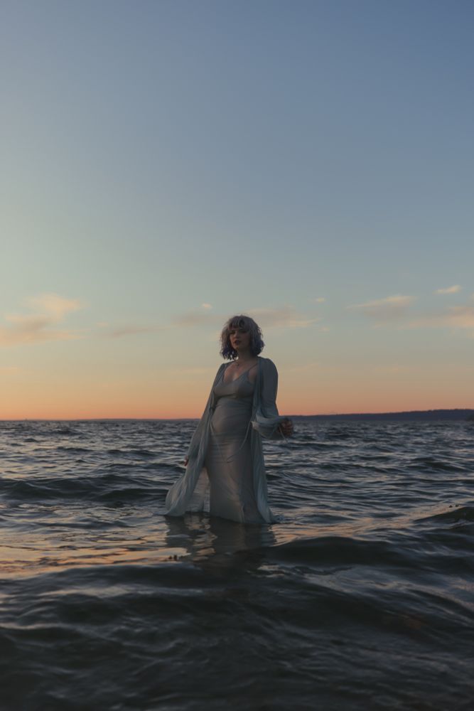 Woman stands in the ocean submerged to the knees wearing a sheer teal dress and sheer floor length robe 