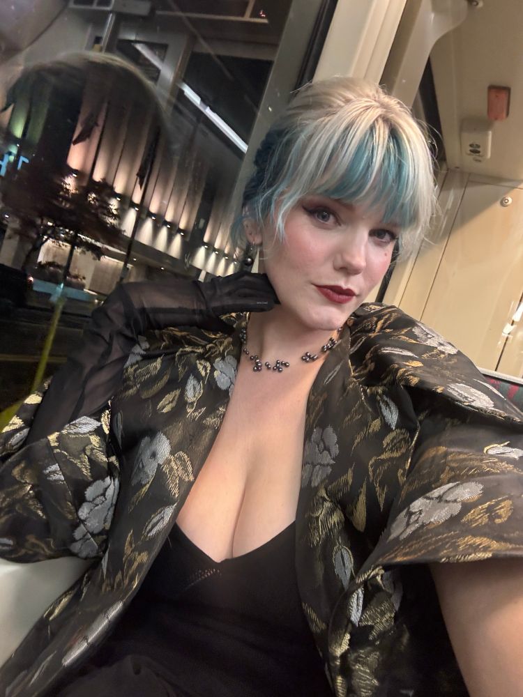 Woman sits and takes a selfie in a silk brocade (?) vintage jacket and low cut black dress, silver and blue hair in an updo, wearing black pearls and red lips