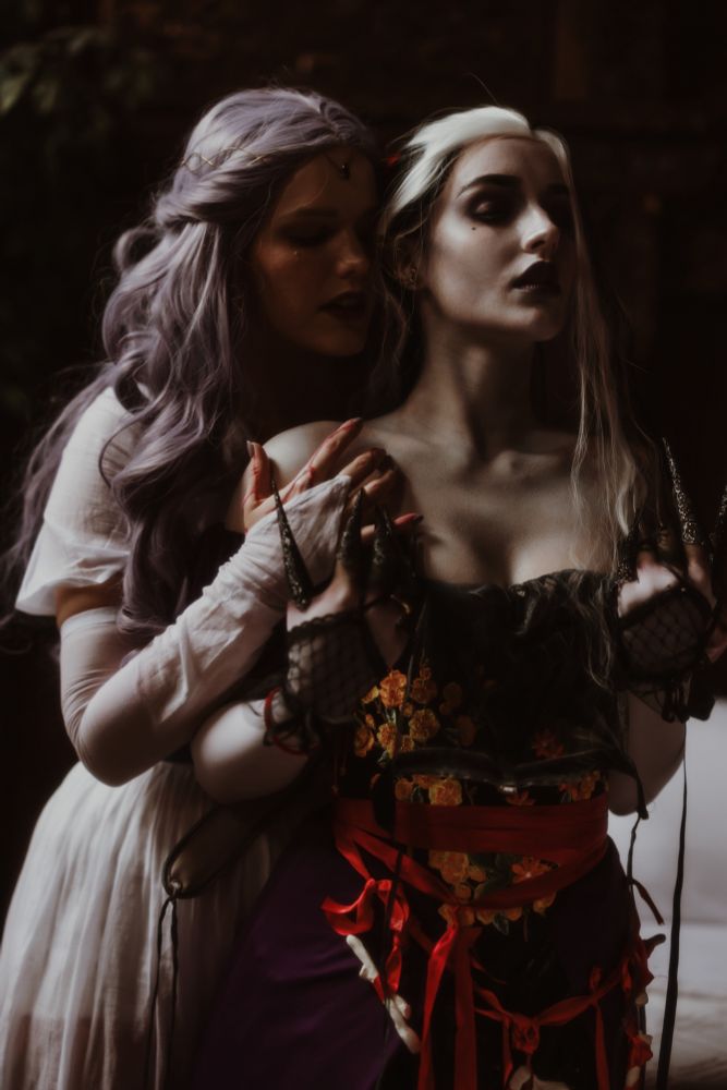Woman with purple hair wearing white dress and gloves stands behind and to the side of woman with pale skin and black and white hair. Purple haired woman is gently touching black haired woman’s bare shoulders