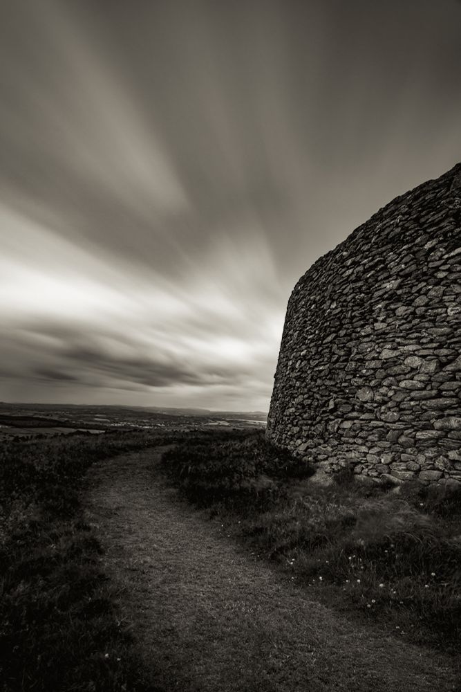 A long exposure shot of a path curving around an ancient ring fort in Ireland. The clouds streak across the sky giving a smooth but epic look and feel to the course, unmovable rocks that make up the barrier wall of this fort. The black and white tones give a timeless feel to the photograph.