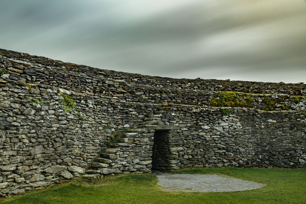 A long exposure shot of the only entrance of an ancient ring fort in Ireland. The clouds streak across the sky giving a smooth but epic look and feel to the course, unmovable rocks that make up the barrier wall of this fort. Stairs run up and down the sides of this fort to access the ramparts, where ferns grow down the sides. 