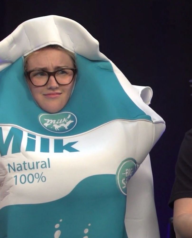 Ashley Johnson wearing a costume of a carton of milk. Her face is peaking through a big hole in the top and that’s all you can see of her. She looks confused and mildly concerned. 
