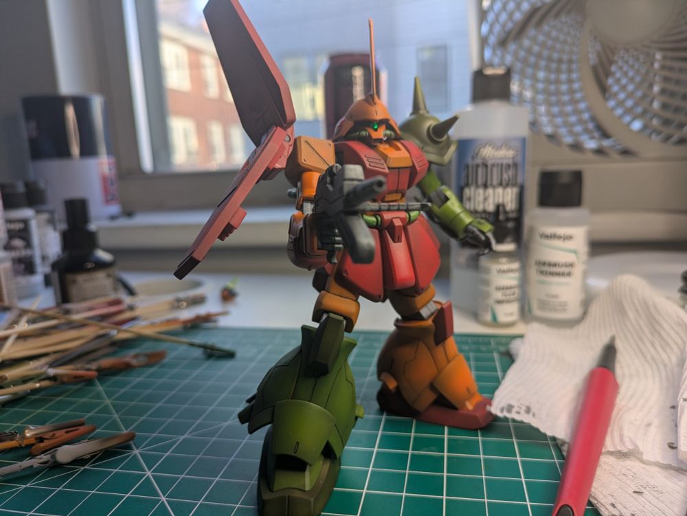 An orange and red mech model from Gundam Zeta holds a beam rifle in its right hand, left arm and right leg are green. A messy workstation is visible around it. It is facing toward the camera.