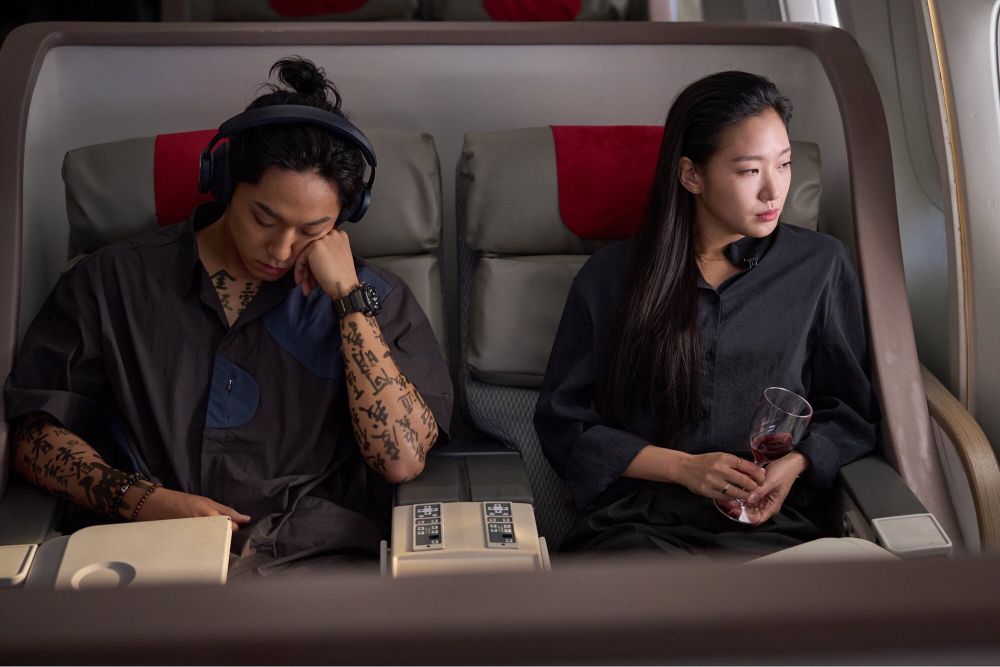 Lee Dohyun and Kim Goeun in a still from the movie Exhuma. Lee Dohyun is wearing the iconic over-ear headphones and a shirt with rolled up sleeves that reveals writing up his arms and you can also see writing on his chest through the open collar of his shirt. His hair is tied up in a small knot. He’s sleeping, face squished into the back of one hand. Kim Goeun is looking out the window, holding a wine glass, gaze set and almost stern. They’re sitting side by side in an airplane, maybe? Both wearing tailored, dark shirts.