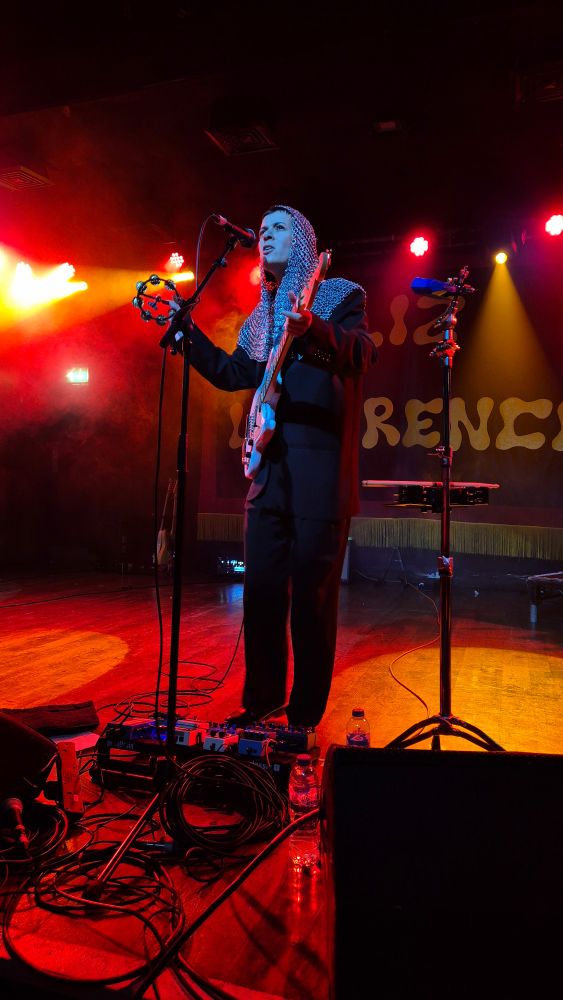 Liz Lawrence on stage wearing a chainmail hood over a black suit. She's singing, shaking some bells and ready to play guitar. 