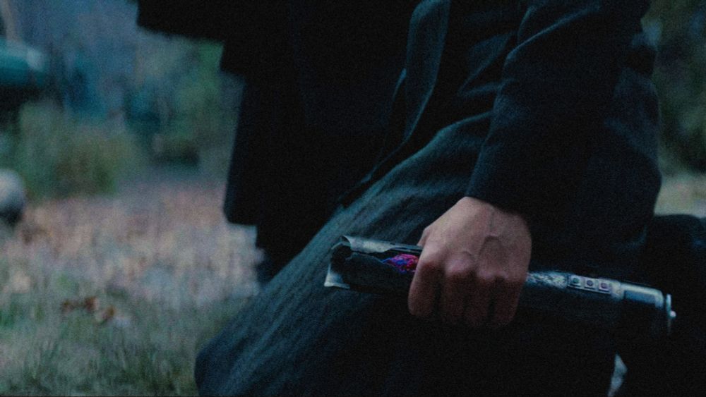 Osha in The Acolyte finale, knelt on the ground on Brendok. Master Sol's lightsaber is the focus, clutched in her hand. The crack shows the blue kyber crystal is turning red. 