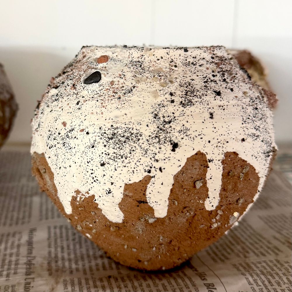 A small moon jar-shaped vessel sits on a board on a wire shelf in front of a plain white wall. It is covered in black textured slip and the top third is  drips of white slip, with a coating of volcanic ash and mica, plus visible spots of sand, rocks, and other textured aggregates visible on the surface. It has not yet been fired so is full of potential.