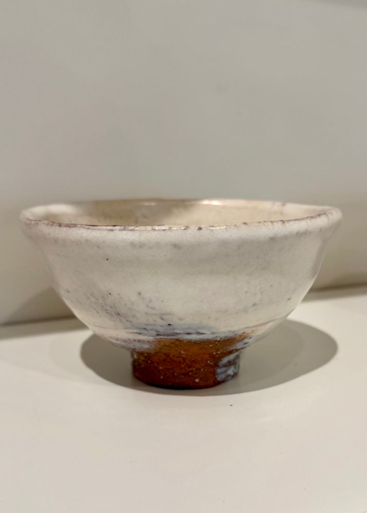 A wide mouthed white glazed Teabowl with groggy red clay visible at the foot sits on a white shelf. 