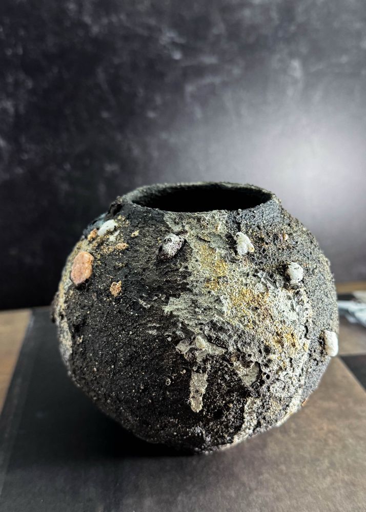 A dark, textured, moon-jar shaped vessel sits on a black book against a black backdrop. Dark slip, sand, rocks, lava stone, and drippy slip are all visible textured on the piece. White stones dot this facade. 