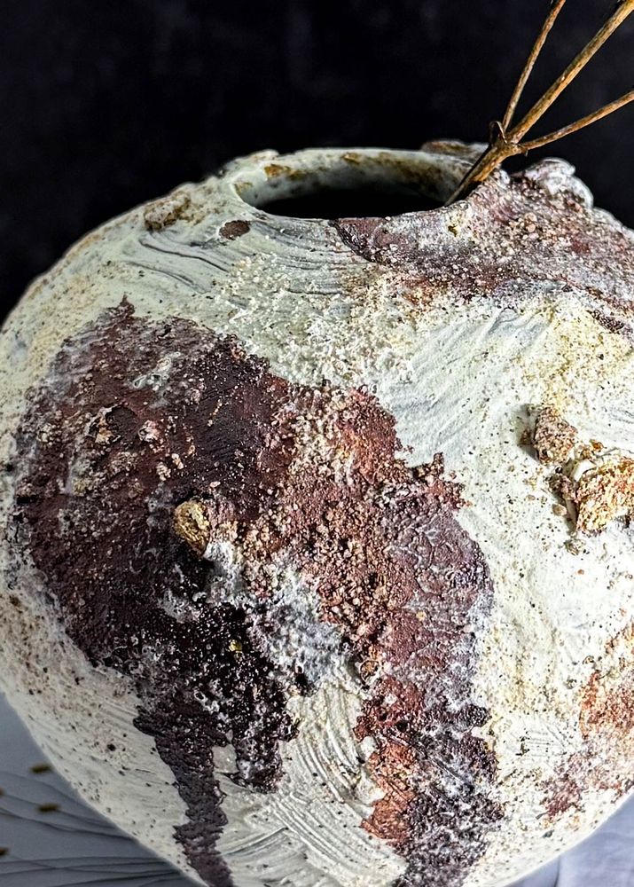 A small orb-shaped vase in close up. White slip brush stroke textures are visible all over with fields of burgundy oxide wash and patches of stones and sand across the surface. It has a small opening perfect for a few buds or dried flowers. 