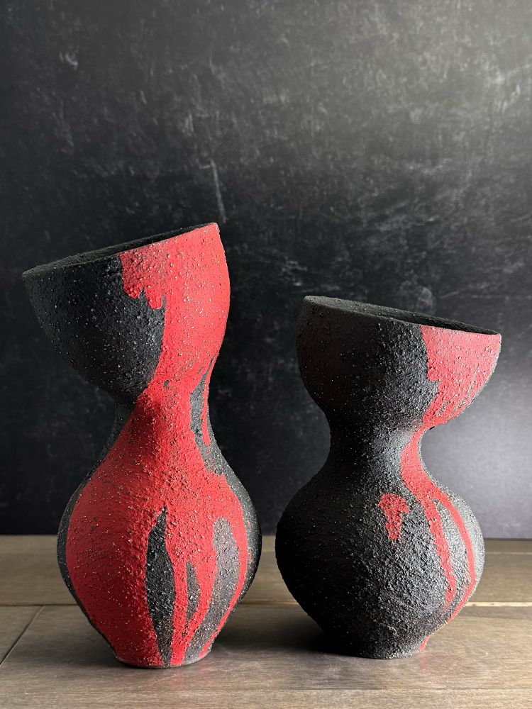 Two vases, the left slightly taller than the right, sit on a wooden table in front of a black marbled wall.  Each vase has an elegant low wide curve that narrows into a skinnier neck before widening back into a trumpet-like opening. They each tilt slightly away from each other. Each is black with heavy texture and drips of bright red running down their sides. I call them “Sway” vases. 