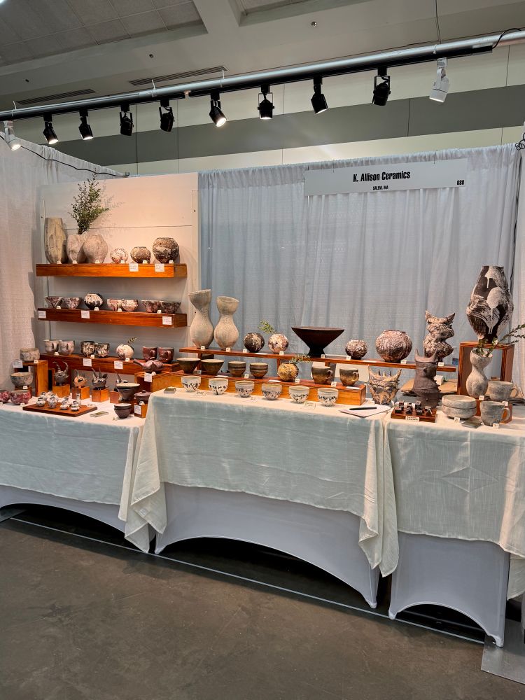 A fine craft fair booth with white curtain, linen-draped tables, and wooden shelf accents displays a large collection of textured, earthy pottery. 