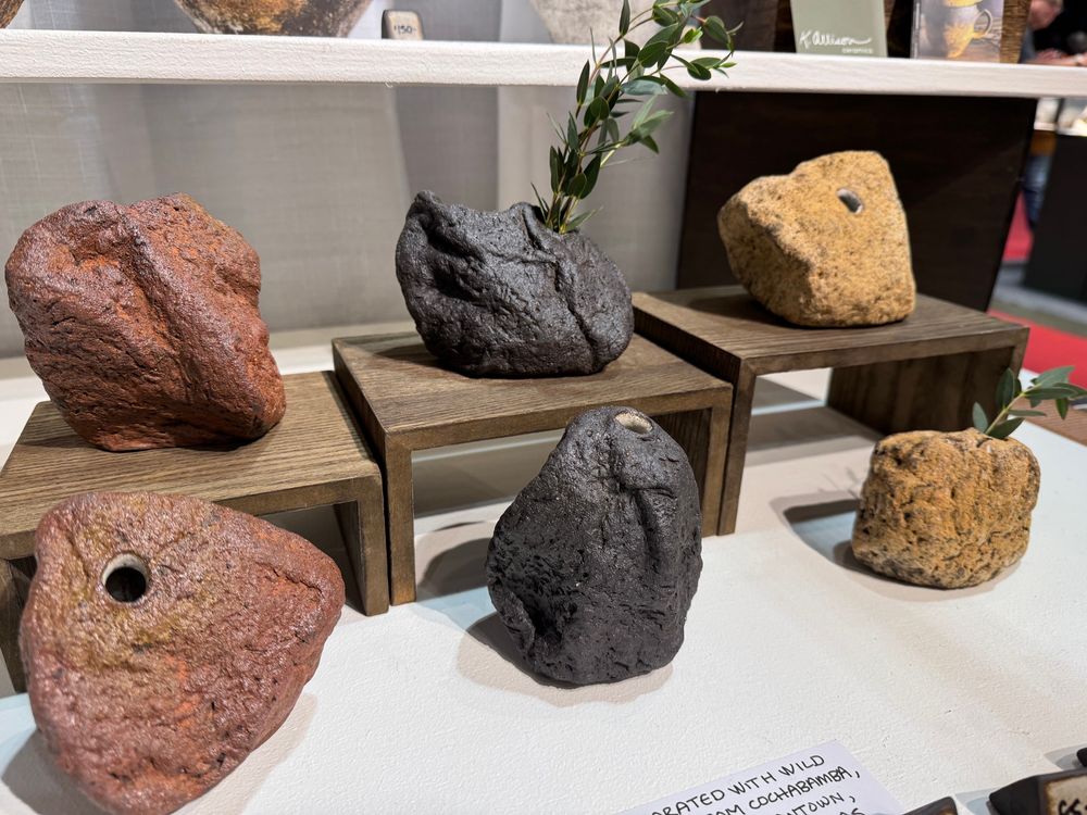 Six bud vases, each shaped and textured like a rock or mountain range. Two are earthy red with a different texture of red wild clay. Two are semi-glossy textured black with red wild clay. Two are an earthy orange-yellow with volcanic ash sprinkled over their texture. Each is a unique shape. 