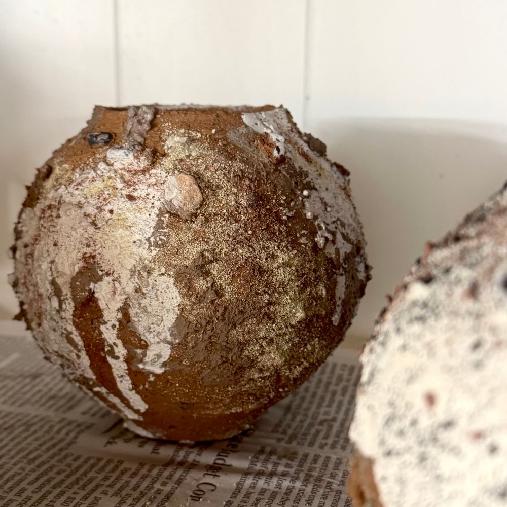 A small moon jar-shaped vessel sits on a board on a wire shelf in front of a plain white wall. It is covered in dark slip and drips of white slip, with spots of sand, rocks, and other textured aggregates visible on the surface. It has not yet been fired so is full of potential.