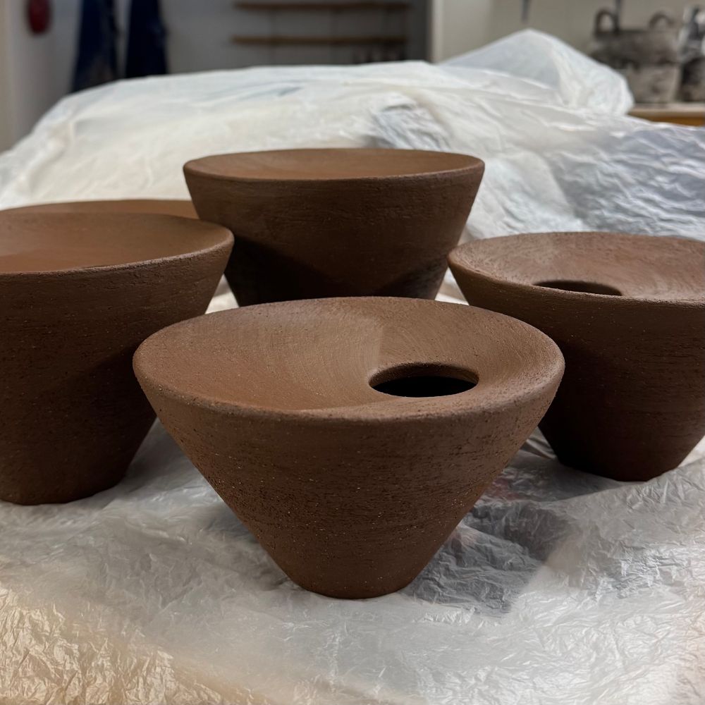 A closeup side profile of greenware dark clay vases in progress sitting on plastic wrapping. A shorter one is shown in full. Three others are partially cropped. They are V shaped with an inward curved top and a small uncentered opening for flowers. 