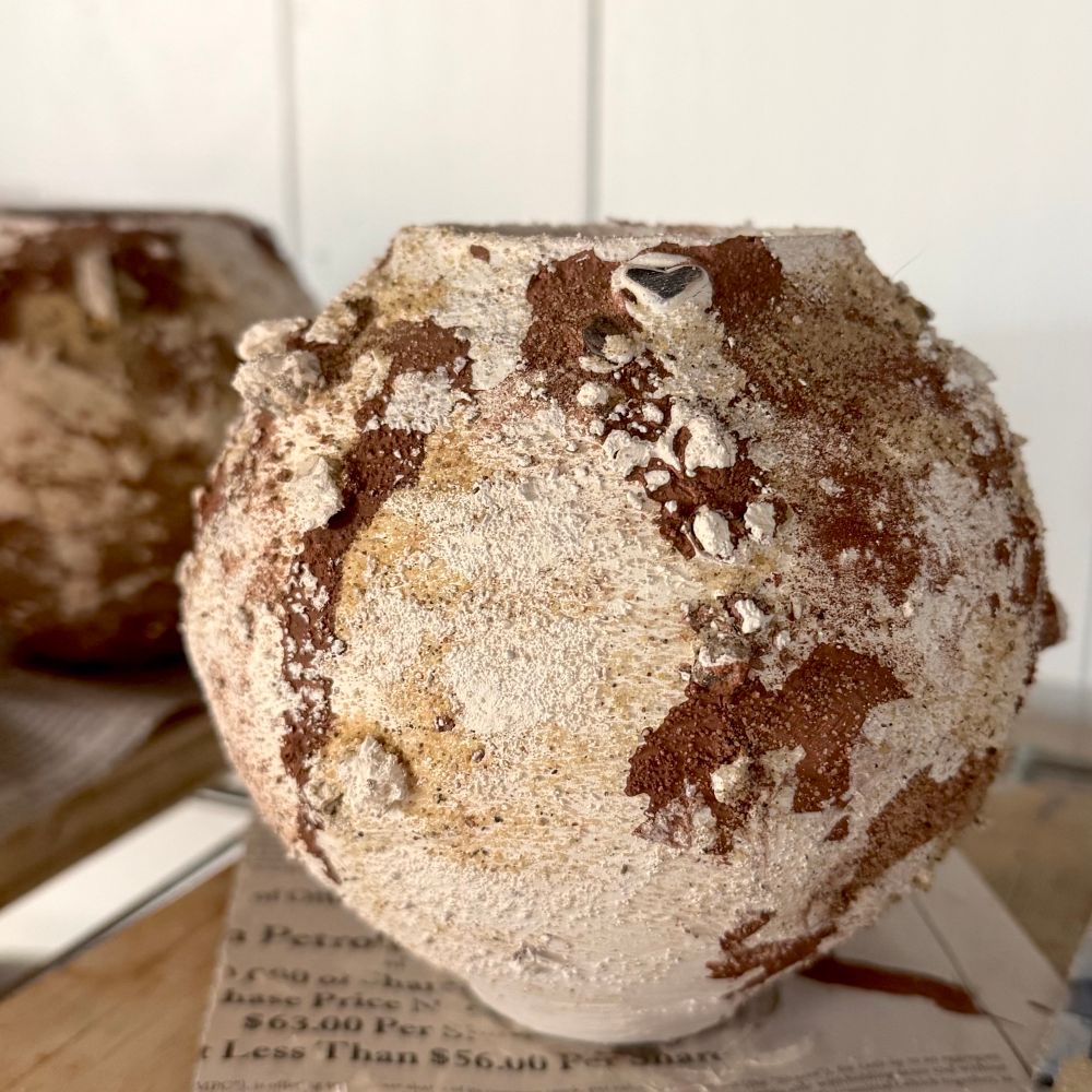 A small moon jar-shaped vessel sits on a board on a wire shelf in front of a plain white wall. It is covered in white slip and drips of burgundy oxide, with spots of sand, rocks, and other textured aggregates visible on the surface. It has not yet been fired so is full of potential.