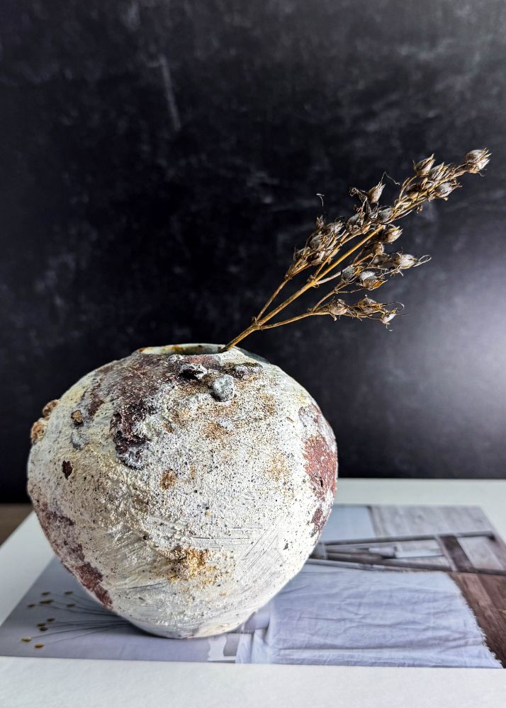 A small orb-shaped vase from its whitest side. White slip brush stroke textures are visible all over with fields of burgundy oxide wash and patches of stones and sand across the surface. It has a small opening perfect for a few buds or dried flowers. 