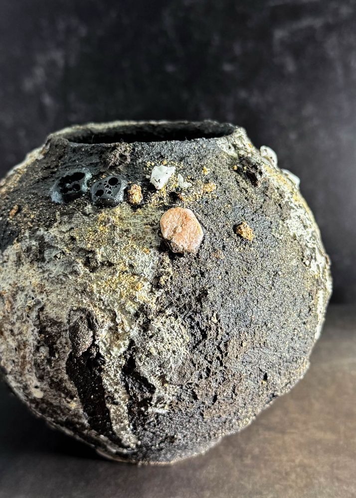 A dark, textured, moon-jar shaped vessel sits on a black book against a black backdrop. Dark slip, sand, rocks, lava stone, and drippy slip are all visible textured on the piece. It also features one almost rounded embedded pink rock that acts as its focal point. 