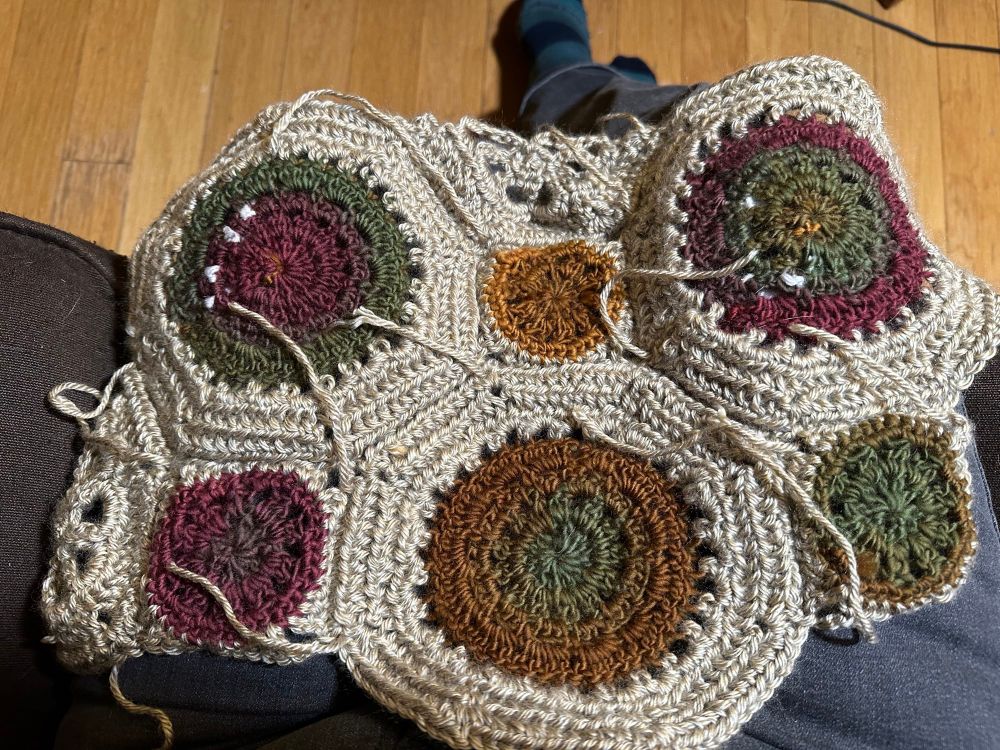 The reverse side of part of a crochet blanket in tan, yellows, greens, and maroon. There are many yarn ends.