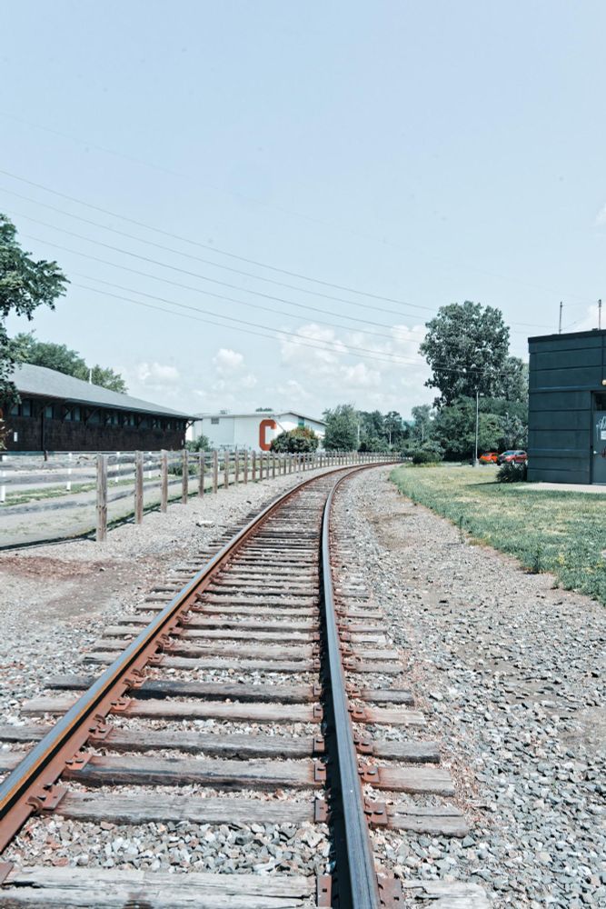 Railroad tracks receed into the background and curve to the right as they go out of sight.  On the left side is a big brown building with a split rail fence in front and behind that a low white building with a red C on it,  on the right a grassy lawn and a little bit of a dark colored building,  some powerlines cross the sky which is blue except for some cumulus clouds in the distant background