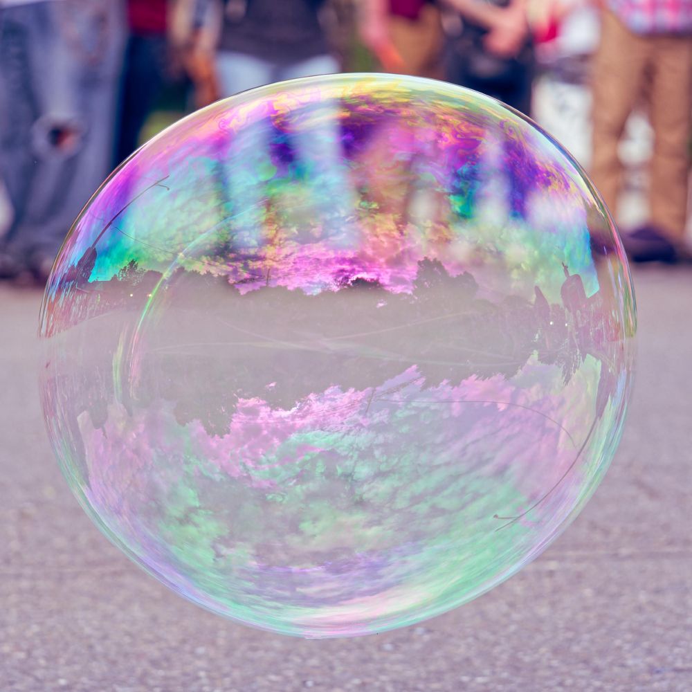 An almost spherical bubble is colored with iridescent reflections above an asphalt road,  behind find a blurred-out crowd where you see people's legs and sometimes folded hands.