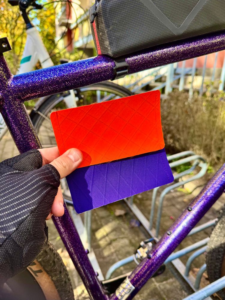 Eine Person mit fingerlosen Fahrradhandschuhen hält ein zweifarbiges Stoffmuster vor den Rahmen eines Fahrrads. Die obere Hälfte des Etuis ist leuchtend orange, die untere Hälfte kräftig violett. Der Fahrradrahmen ist auffällig violett mit Glitzereffekt lackiert. Auf dem Oberrohr des Rahmens ist eine schwarze Rahmentasche befestigt. Im Hintergrund sind ein weiteres Fahrrad und ein Fahrradständer zu sehen. Die Szene spielt sich draußen bei sonnigem Wetter ab.