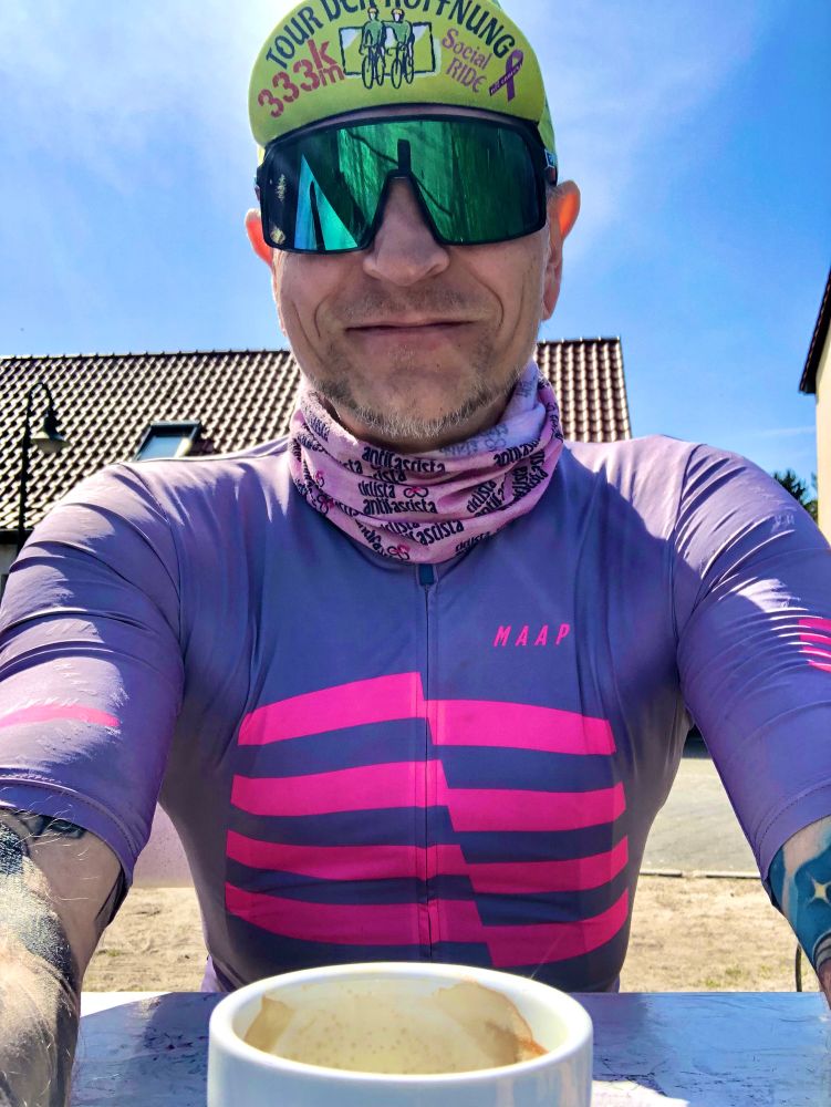 Ein Radfahrer in violett-pinkem Trikot mit der Aufschrift „MAAP“ macht ein Selfie bei sonnigem Wetter. Er trägt eine grüne Mütze mit dem Text „Tour der Hoffnung 333K Social Ride“, eine reflektierende Sportbrille und ein Halstuch mit „antifascista“-Aufdruck. Im Vordergrund steht eine halbleere Tasse Kaffee.