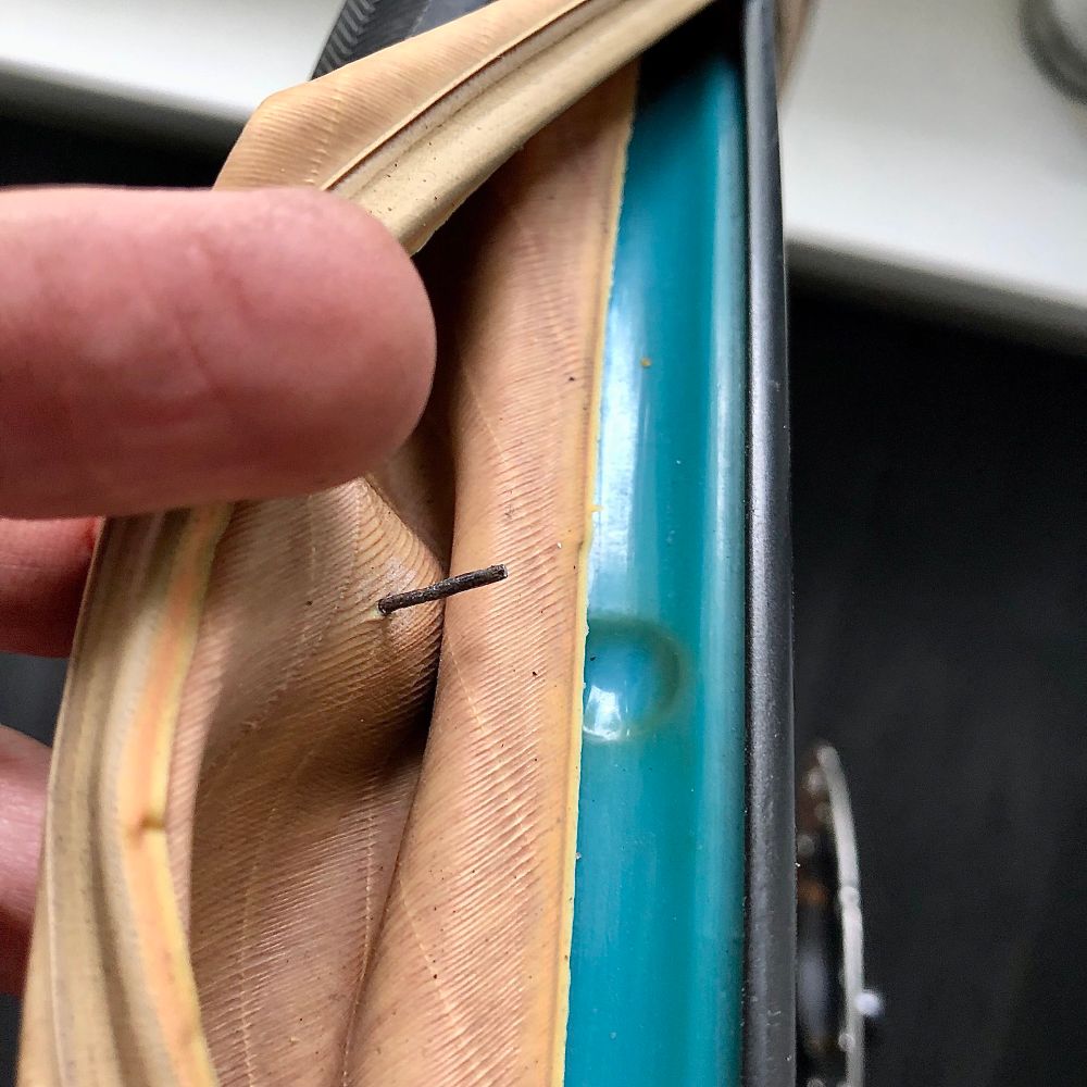Detailaufnahme eines abgezogenen Fahrradreifens, bei dem ein Finger den beige-braunen Mantel zur Seite zieht, um einen eingedrungenen Fremdkörper zu zeigen. Ein dünner Draht oder Metallstück (12mm lang) steckt deutlich sichtbar durch die Innenseite des Reifens. Darunter ist das türkisfarbene Felgenband zu erkennen.