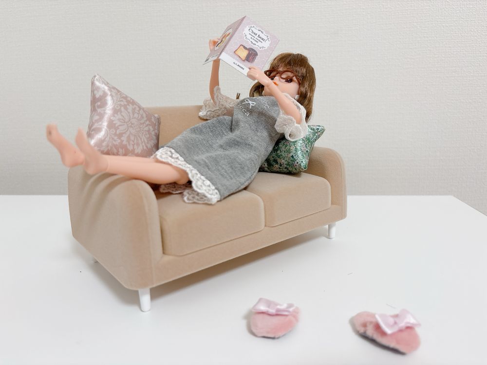 写真：ミニチュアソファに仰向けで寝転がって本を読むリカちゃん人形。ソファ近くにスリッパが散乱している。