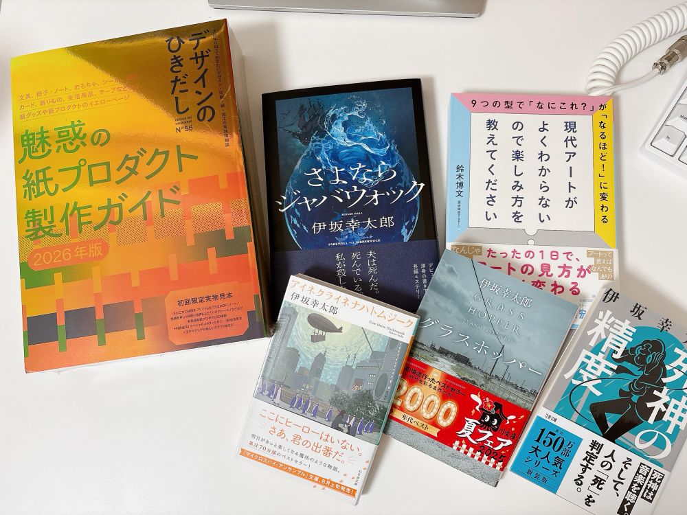 写真：机の上に並べられた数冊の本の写真。『魅惑の紙プロダクト製作ガイド 2026年版』をはじめ、伊坂幸太郎の小説『さよならシナプスウォッグ』『グラスホッパー』『アイネクライネナハトムジーク』『死神の精度』、そして現代アートの楽しみ方を解説する本が置かれている。