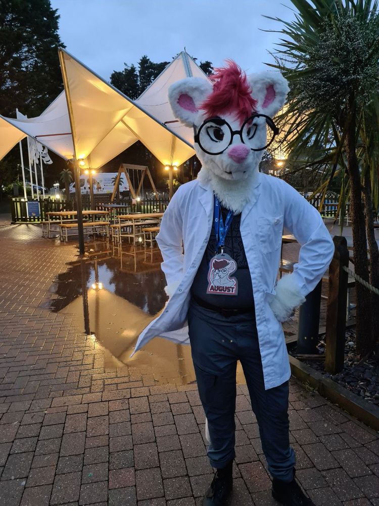 A rat fursuit in a lab coat with big glasses poses hand on hip in front of a large puddle next to outdoor covered seating area at night.