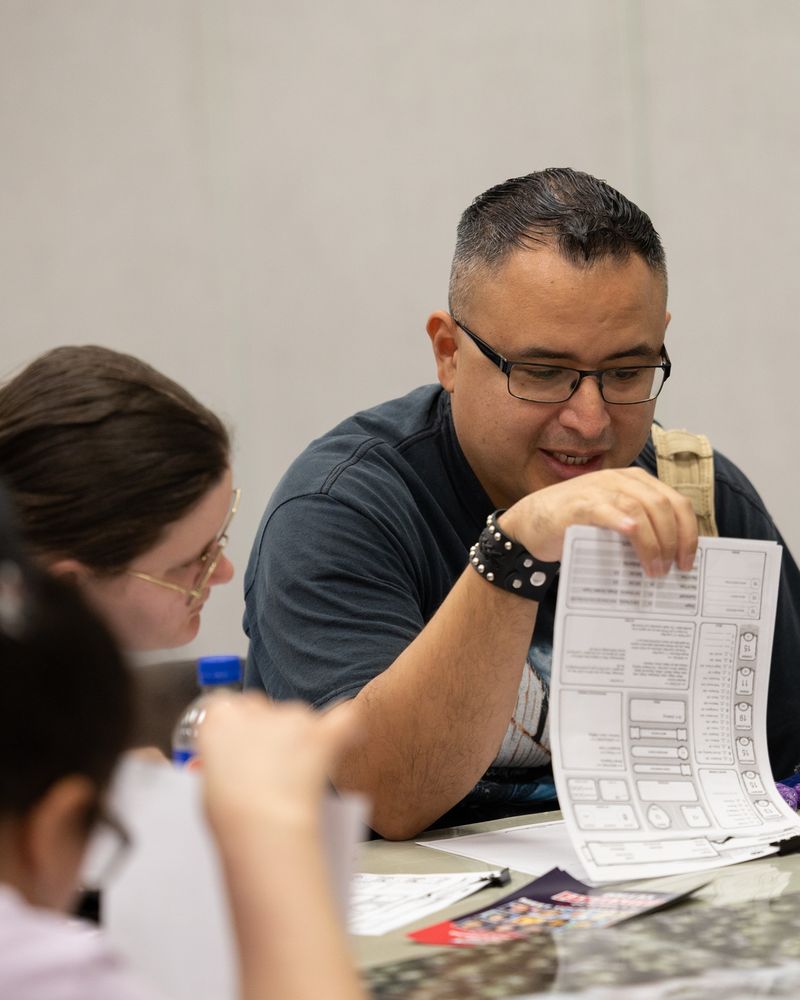 A man with short dark hair and glasses studies a character sheet during a tabletop role-playing session. He holds the sheet up while speaking to others at the table. A woman beside him leans in to look at the page, and various papers, pencils, and small game items are spread out in front of them.